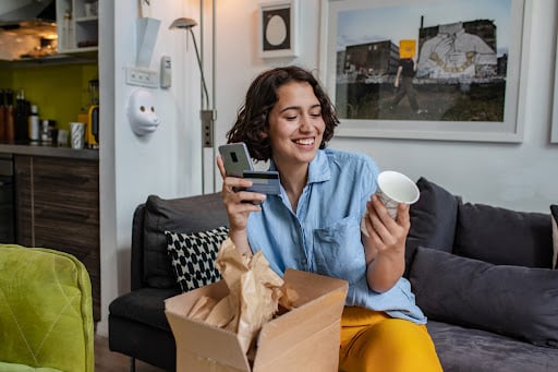 Young woman holding a cup, phone, and credit card as she unpacks the package she ordered online